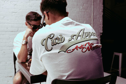 White Donk Tee with "Curb Appeal Art in Motion" design worn by a seated person.