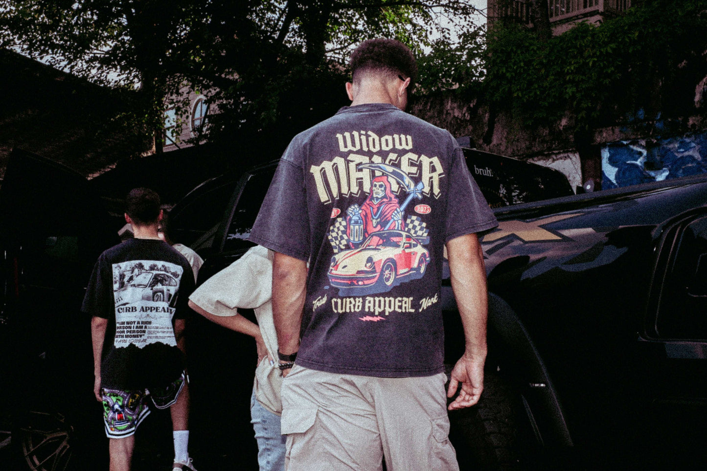 Widow Maker Tee with bold automotive graphics, worn by a model outdoors near vehicles.