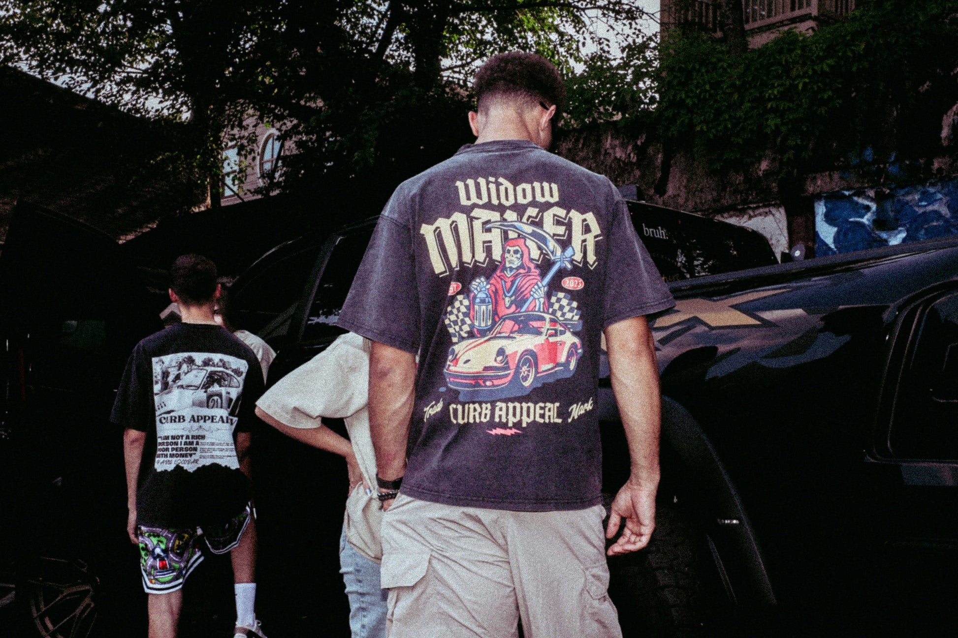 Widow Maker Tee with bold automotive graphics, worn by a model outdoors near vehicles.